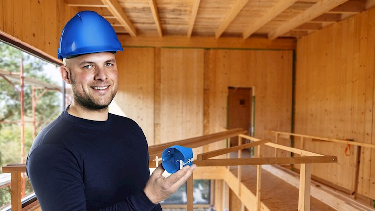 Installateur met blauwe helm houdt een blauwe CLT-inbouwdoos vast in een gebouw van massief hout